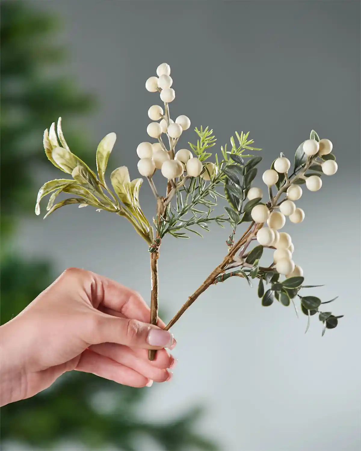 Hand holding a decorative branch with green leaves and white berries against a blurred grey background
