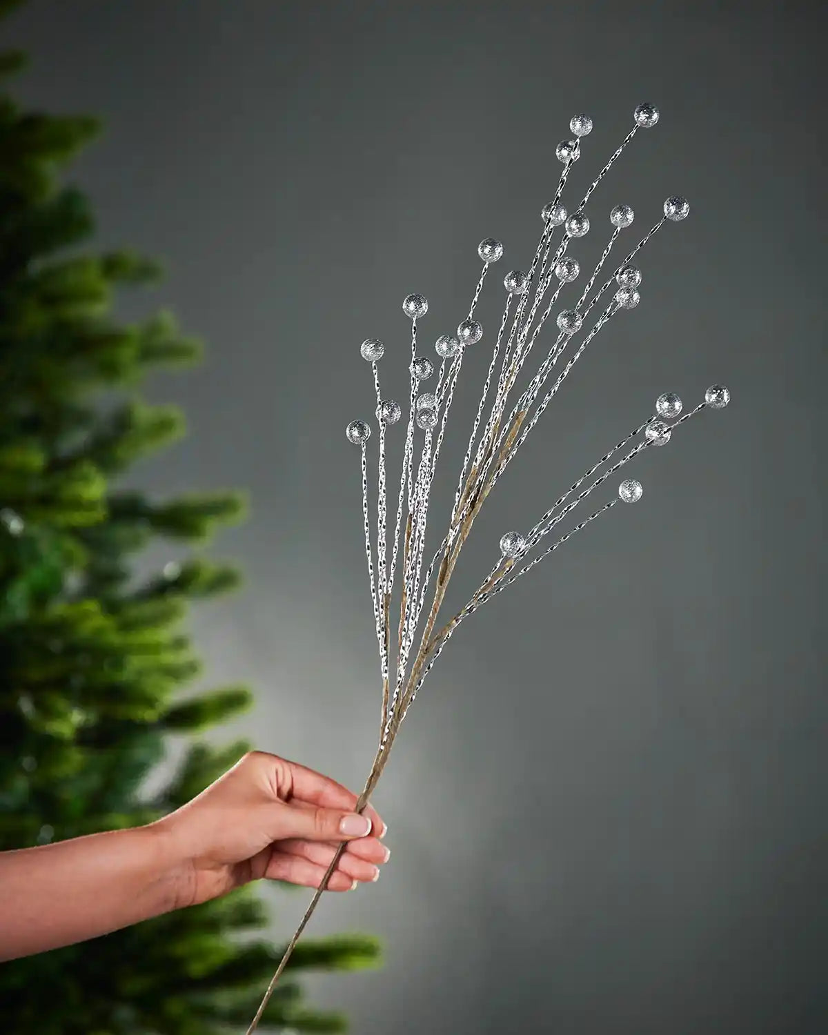 Hand holding a decorative silver tree spray with glitter silver berries on a grey blurry background with green Christmas tree