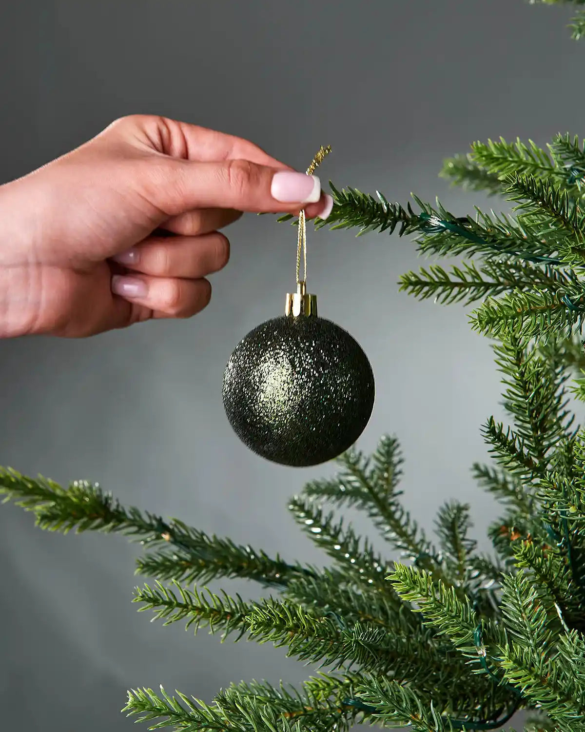 Hand holding a glittery green Christmas bauble on a tree branch against a grey background
