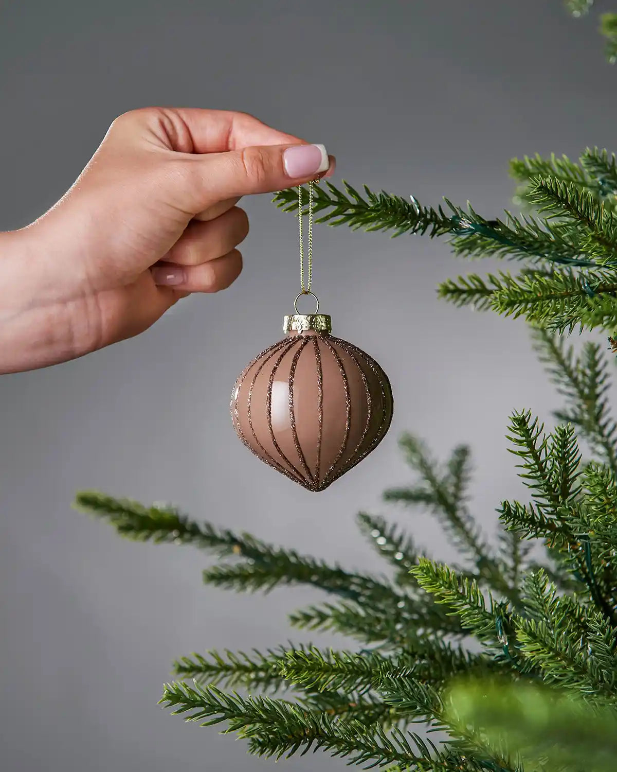 Hand holding a ribbed Latte Christmas bauble against a grey background with green tree branches.