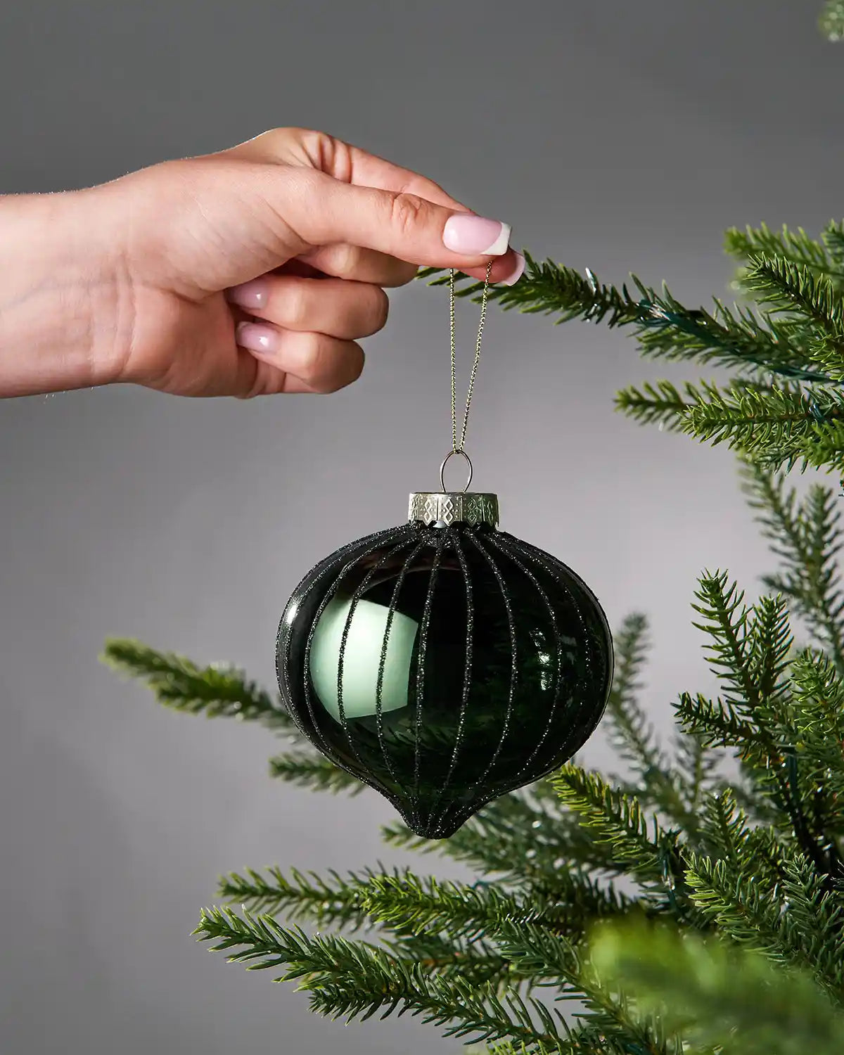 Hand holding a sage green glass ornament on a branch against a grey background