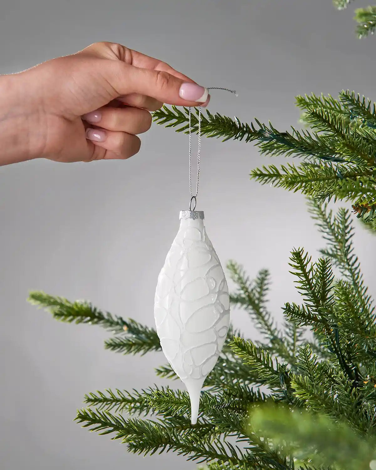 Hand holding a white textured Christmas bauble on a tree branch with a grey background