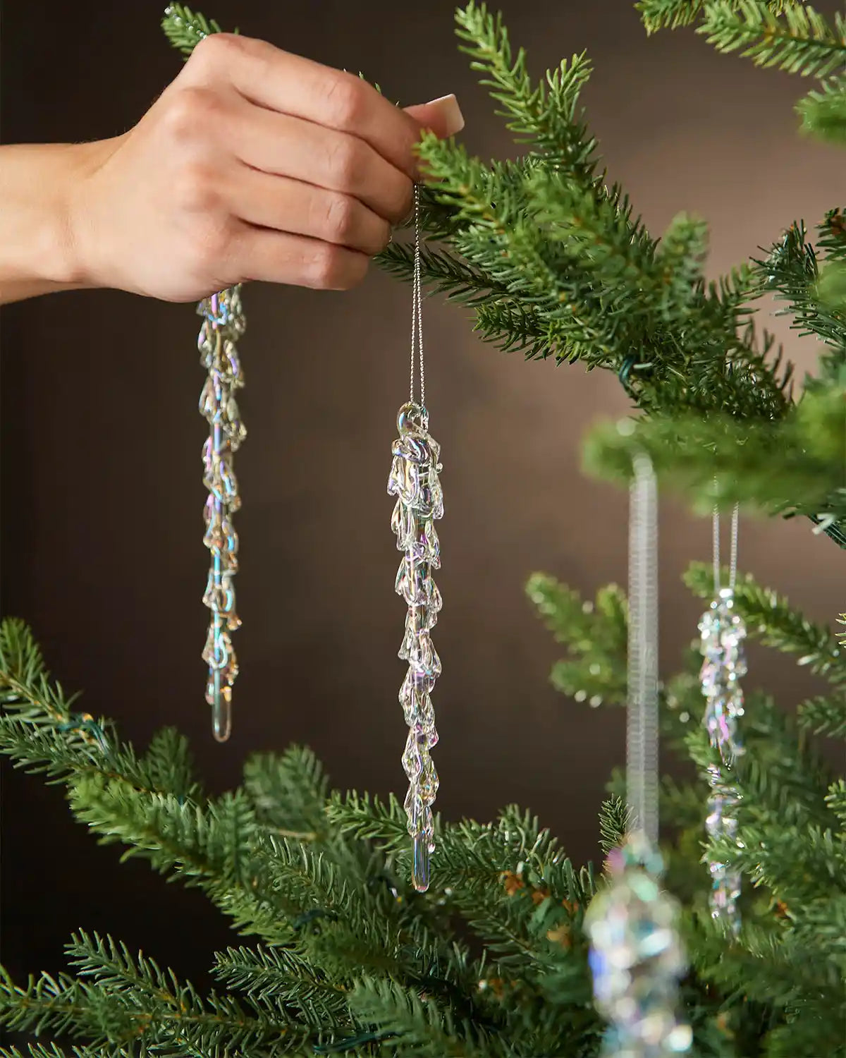 Hand holding an iridescent crystal icicle ornaments hanging from a Christmas tree branch against a brown background 