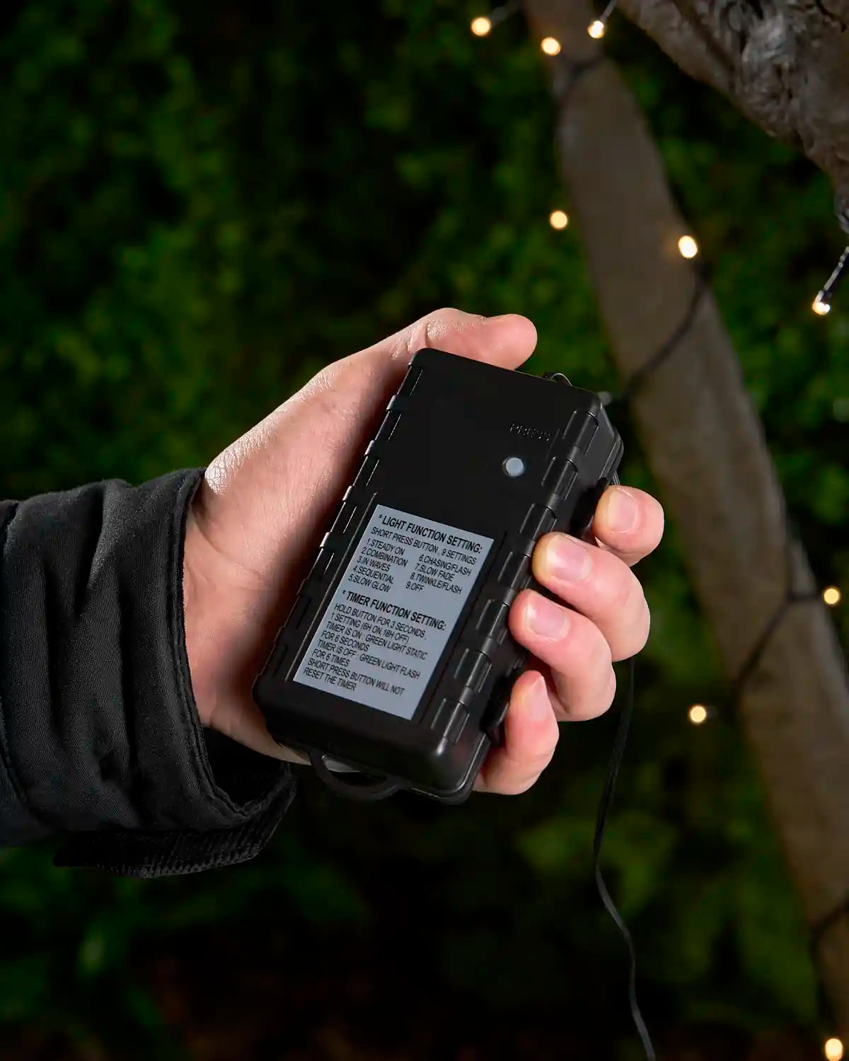 Hand holding black 3C Battery Box with warm white LEDs attached to tree  blurred in the background