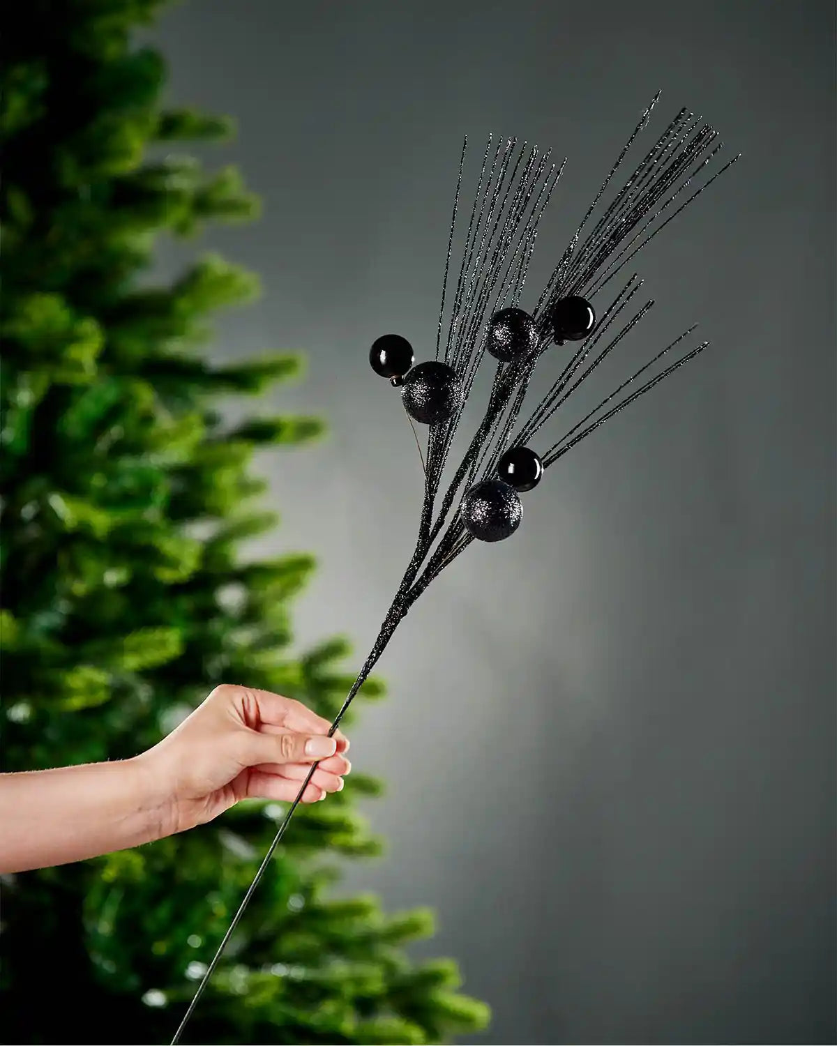 A hand holding the stem of black glitter bauble spray with portion of Christmas tree and grey wall in background