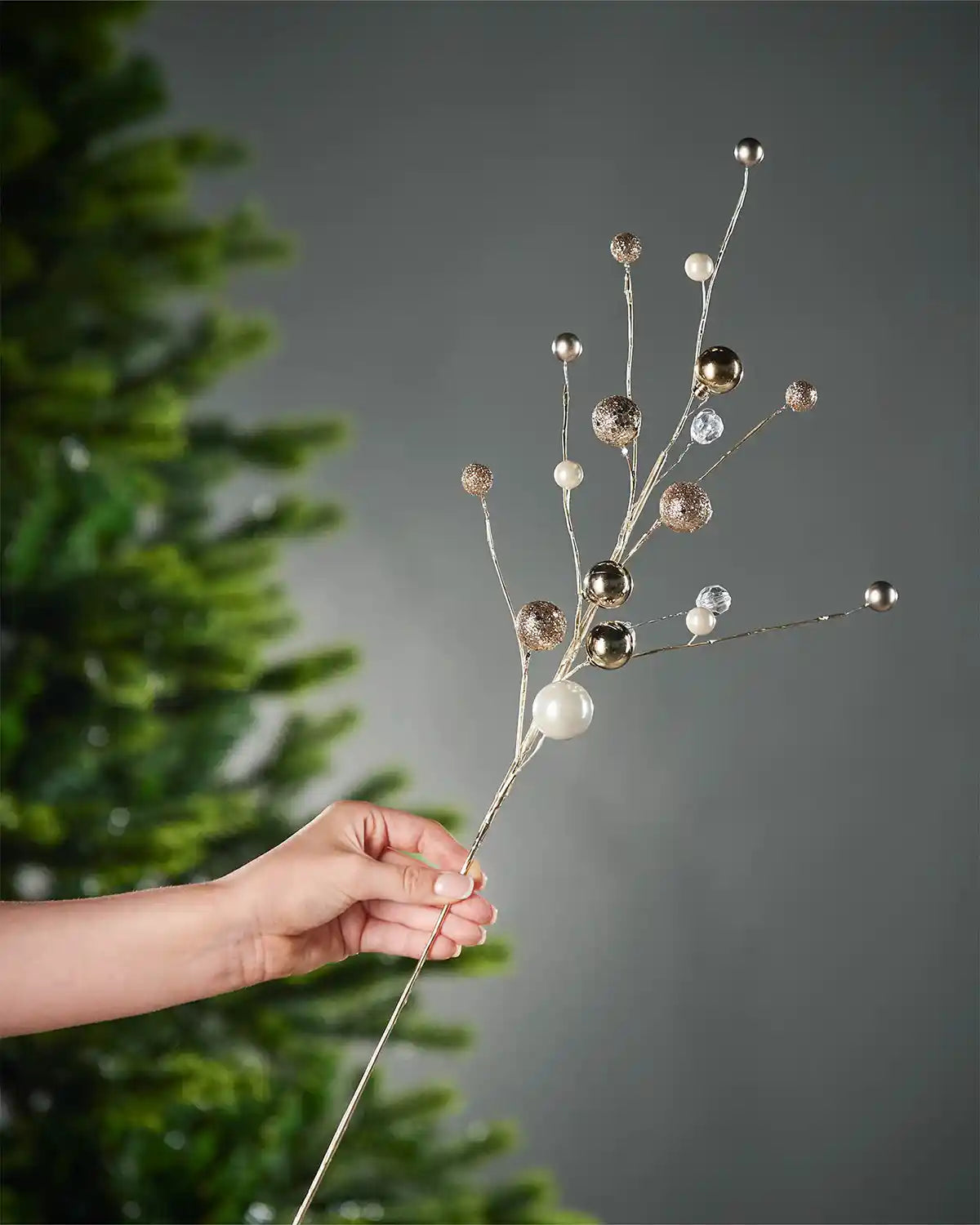 A hand holding the gold bauble and jewelled spray stem in front of Christmas tree and grey wall background