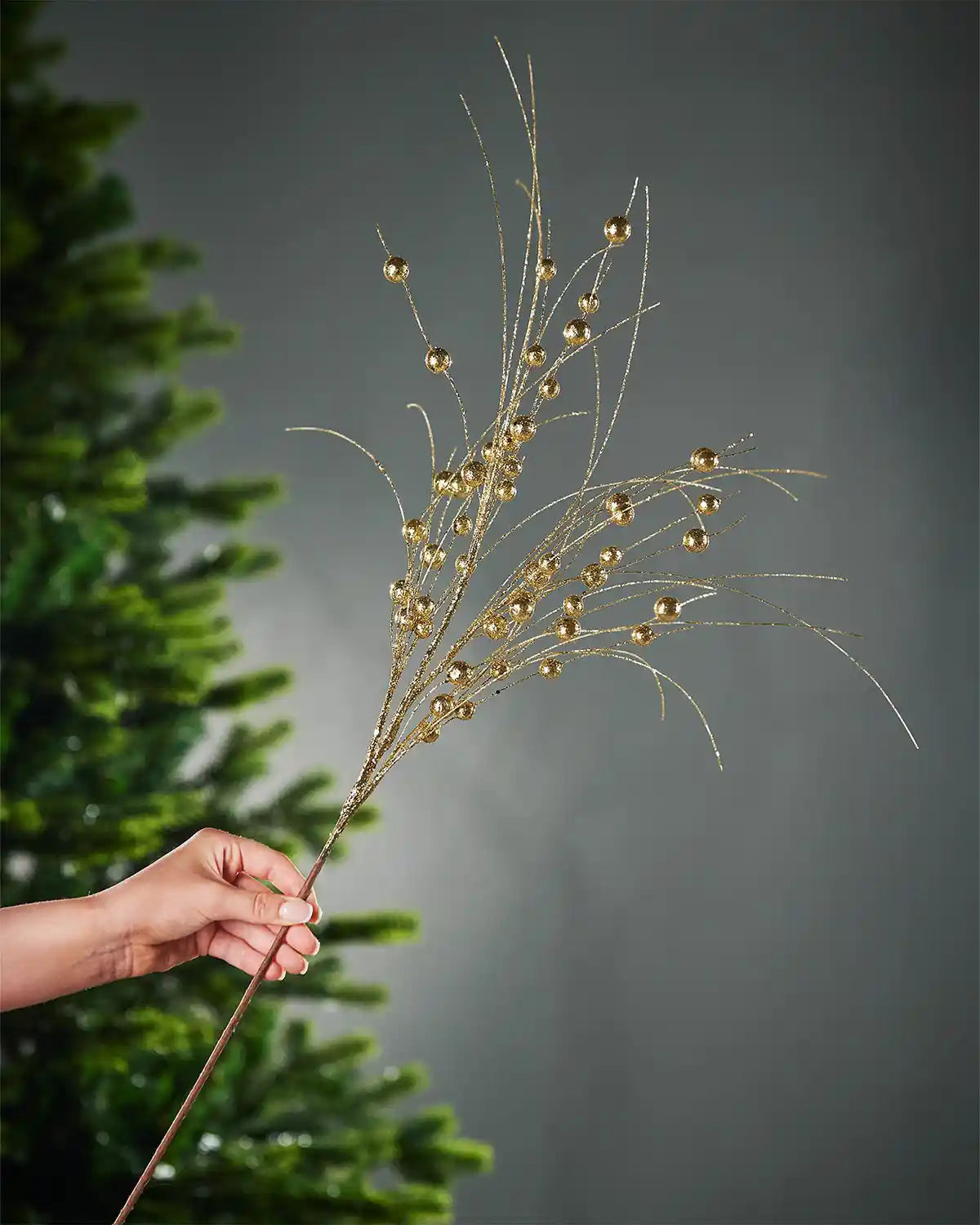 A hand holding the stem of the gold glitter berry spray in front of a Christmas tree and grey wall background