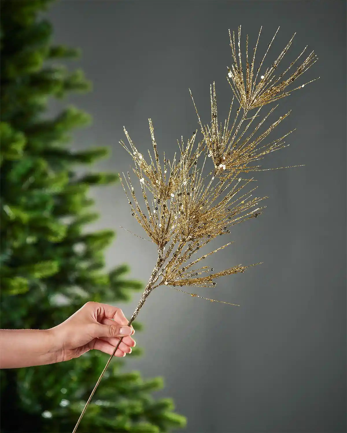 A hand holding the stem of gold glitter firework spray in front of portion of Christmas tree and grey wall