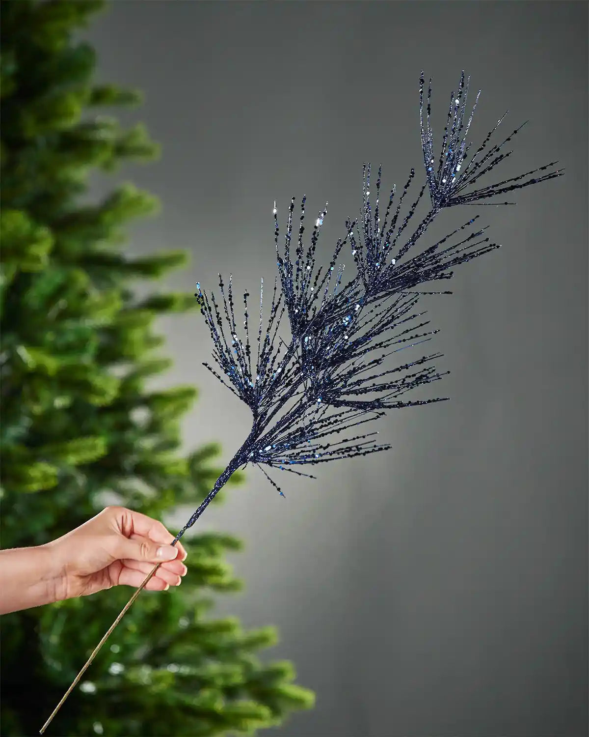 A hand holding navy blue glitter firework spray stem in front of a background of a Christmas tree and grey wall