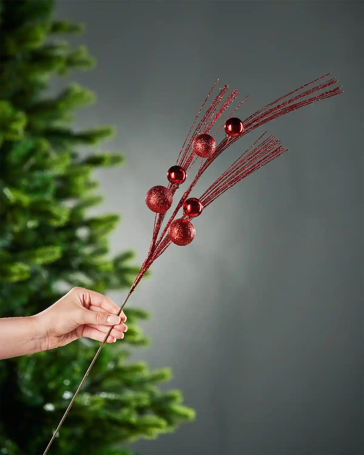 A hand holding the brown stem of the red glitter bauble spray in front of background containing portion of Christmas tree and grey wall