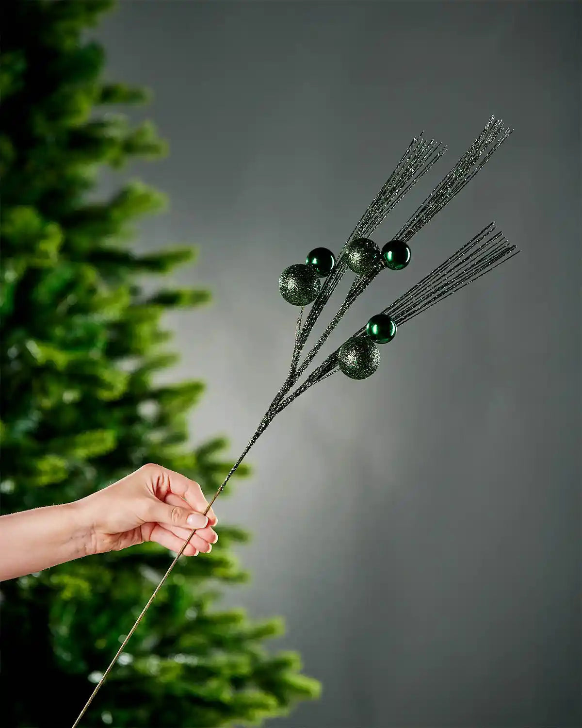 A hand holding sage green glitter bauble spray by the stem with portion of Christmas tree and grey wall in background