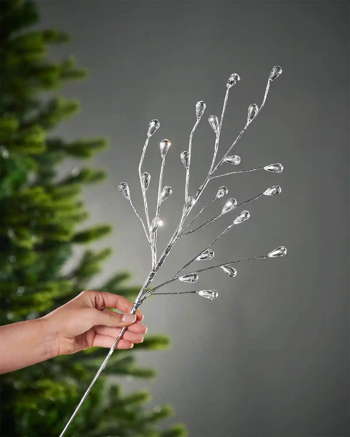 A hand holding silver crystal teardrop spray in front of Christmas tree and grey wall background