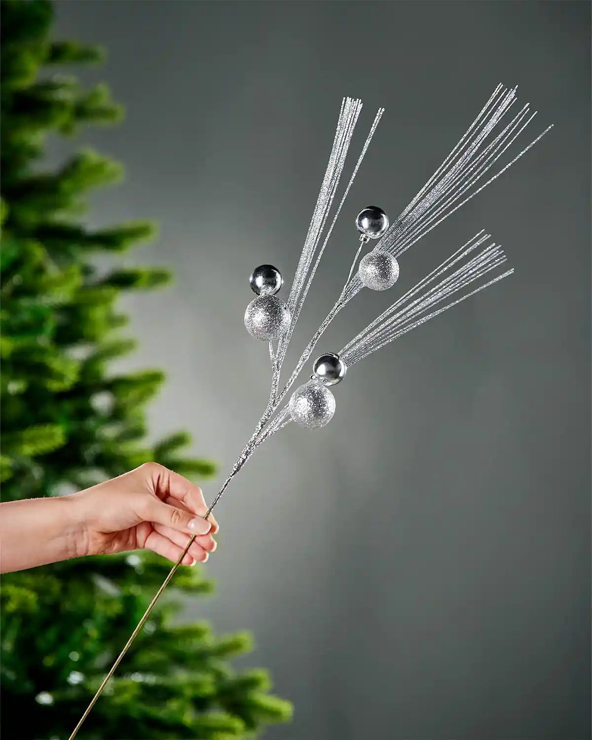 A hand holding the stem of silver glitter bauble spray with portion of Christmas tree and grey wall in background