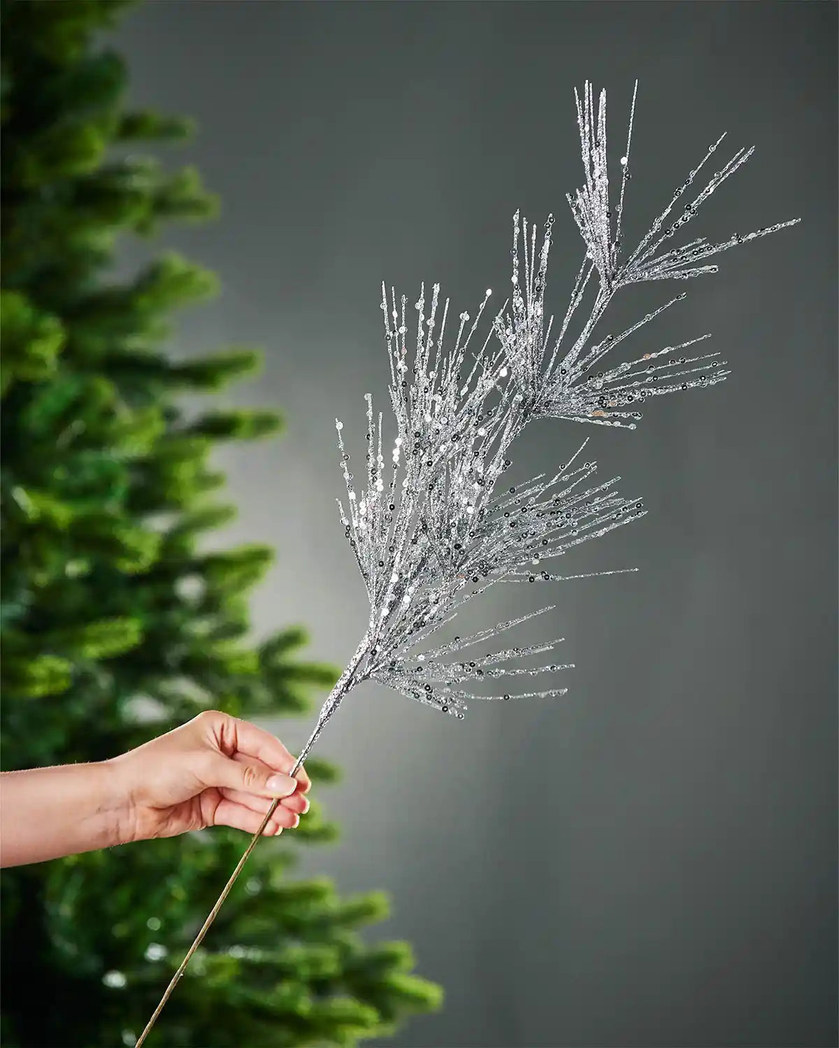 A hand holding silver glitter firework spray stem in front of portion of Christmas tree and grey wall 