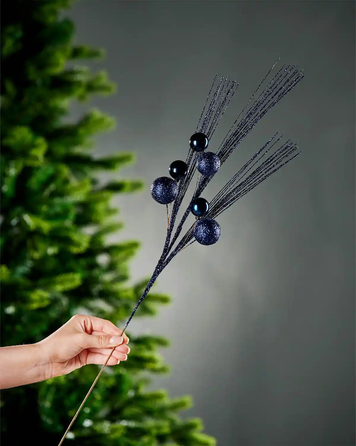 A hand holding the stem of navy blue glitter bauble spray with portion of Christmas tree and grey wall in background