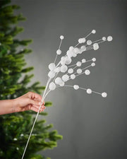 A hand holding the stem of white snowball spray in front of Christmas tree and grey wall background