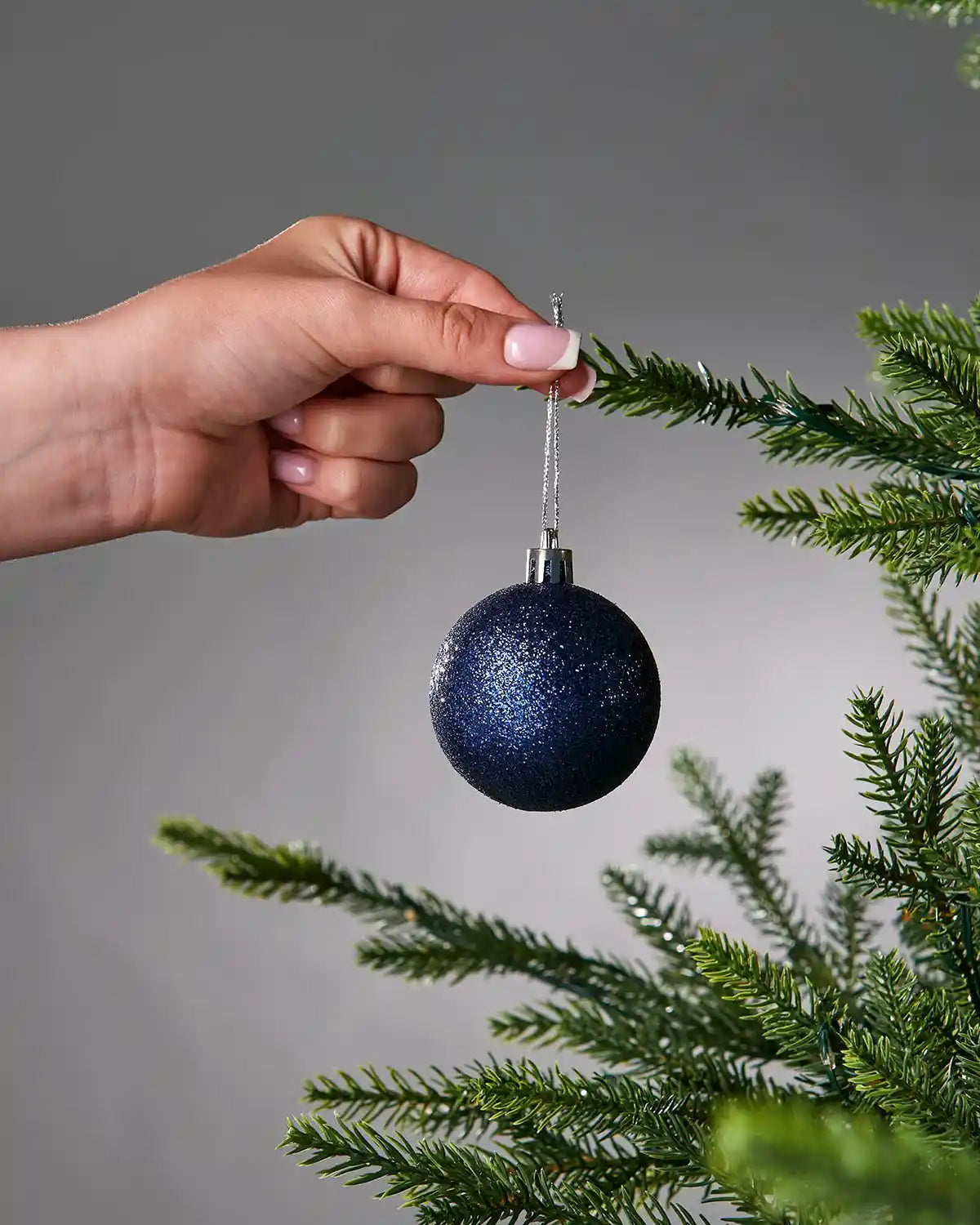 Hand placing a blue glittery Christmas bauble on a tree branch against a grey background