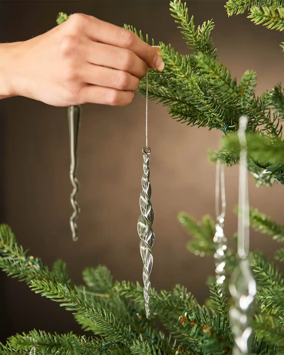 Hand placing a glass icicle Christmas ornament on a tree branch against a brown background