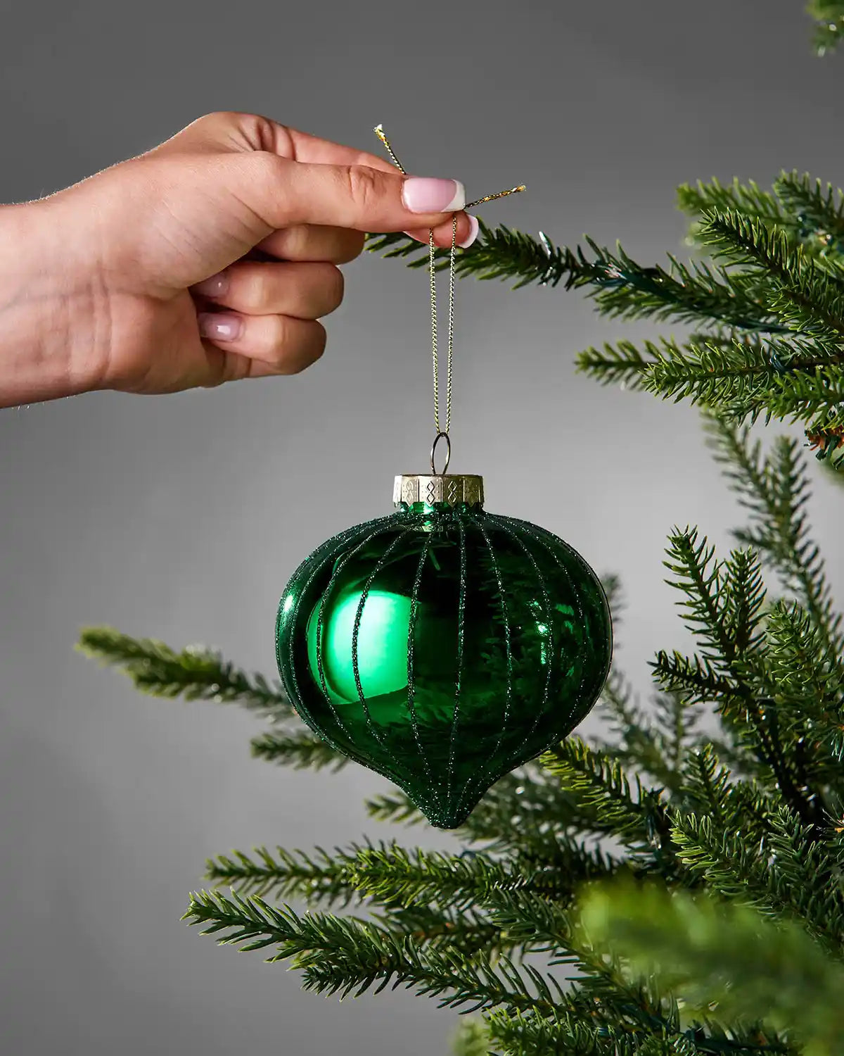 Hand placing a green glass ornament on a tree branch against a grey background