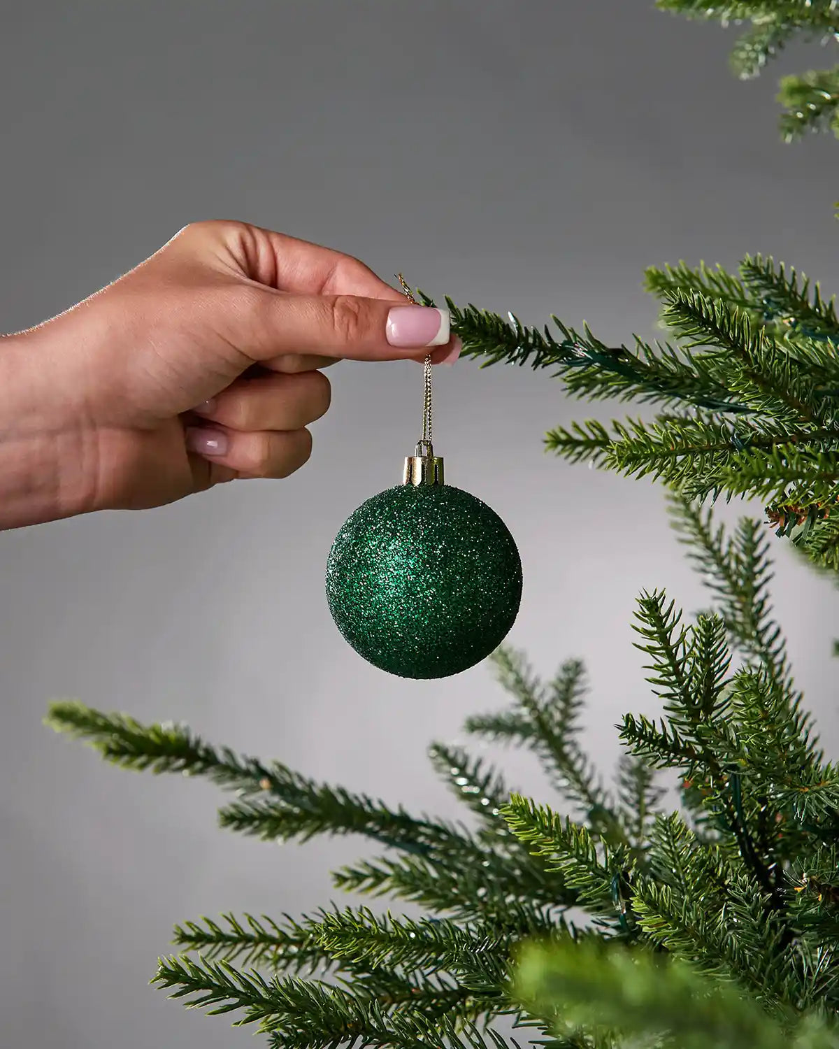 Hand placing a green glittery Christmas ornament on a tree branch against a grey background