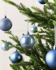 Ice Blue Christmas tree ornaments hanging on green pine branches against a light neutral background.