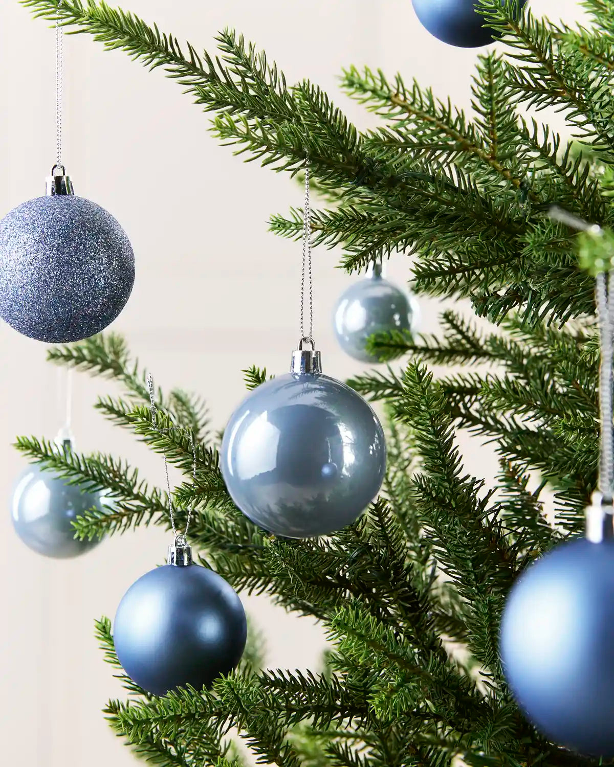 Ice Blue Christmas tree ornaments hanging on green pine branches against a light neutral background.
