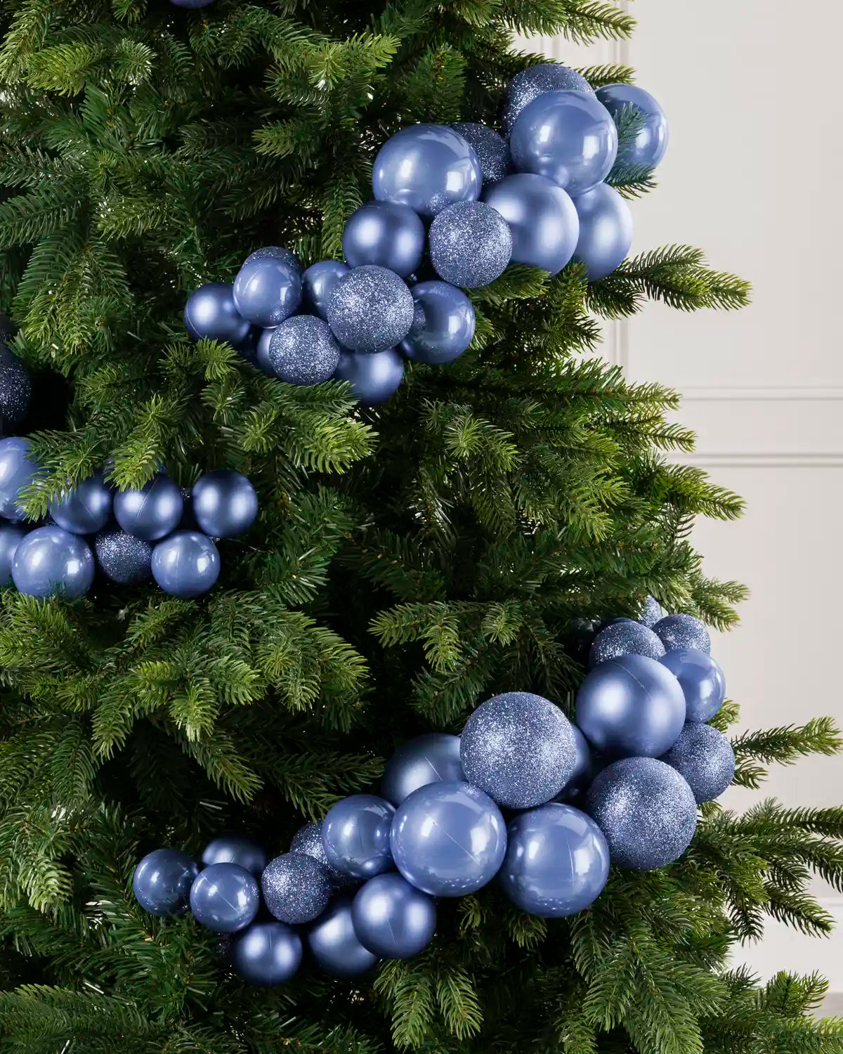 Ice Blue Christmas tree ornaments in glossy and glittery finishes, arranged on a green artificial pine tree against a light neutral wall.