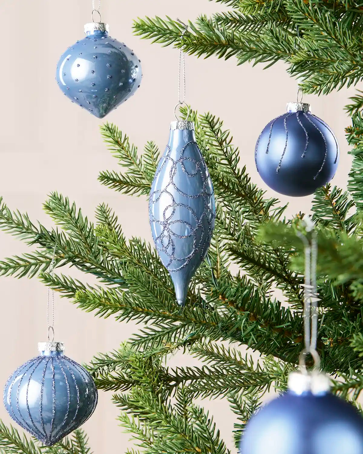 Ice blue glass baubles hanging from Christmas tree branches in teardrop, onion and classic round shapes with a blurry cream background.