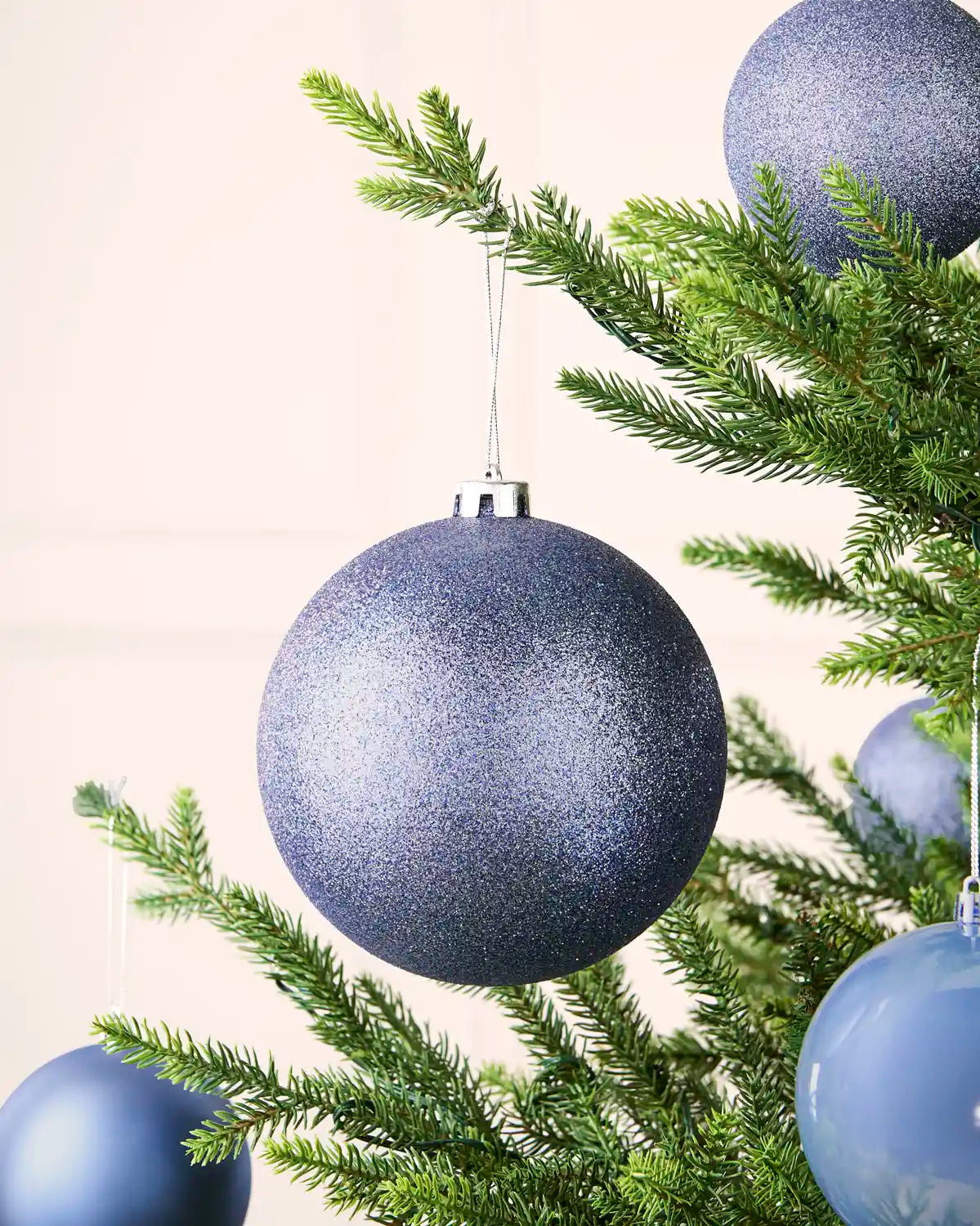 Ice blue glitter oversized bauble with smaller baubles hanging on Christmas tree against a cream background