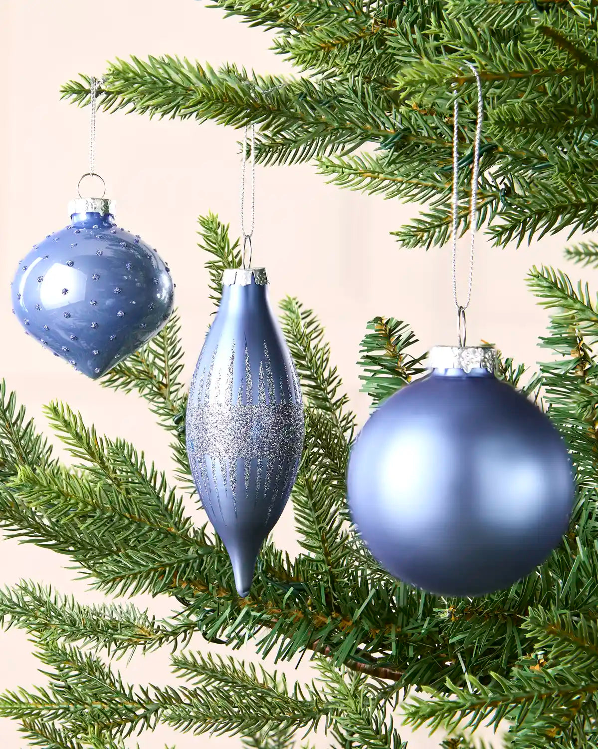 Ice blue hanging baubles in 3 shapes and sizes on a Christmas tree branch against a cream wall 
