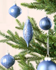 Ice blue hanging baubles in various shapes and sizes on a Christmas tree branch against a cream wall 