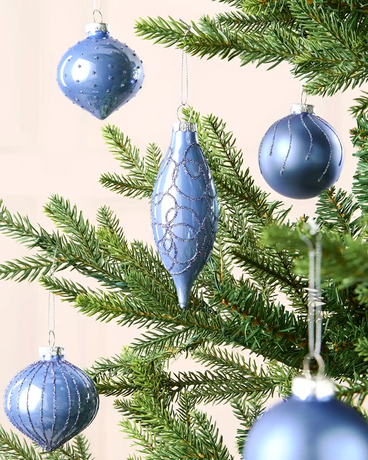 Ice blue hanging baubles in various shapes and sizes on a Christmas tree branch against a cream wall 