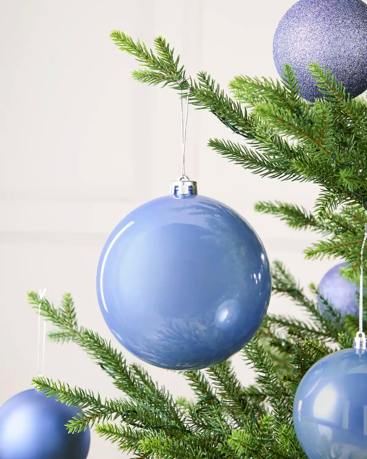 Ice blue shiny oversized bauble with smaller baubles in the background hanging on a Christmas tree against a cream background