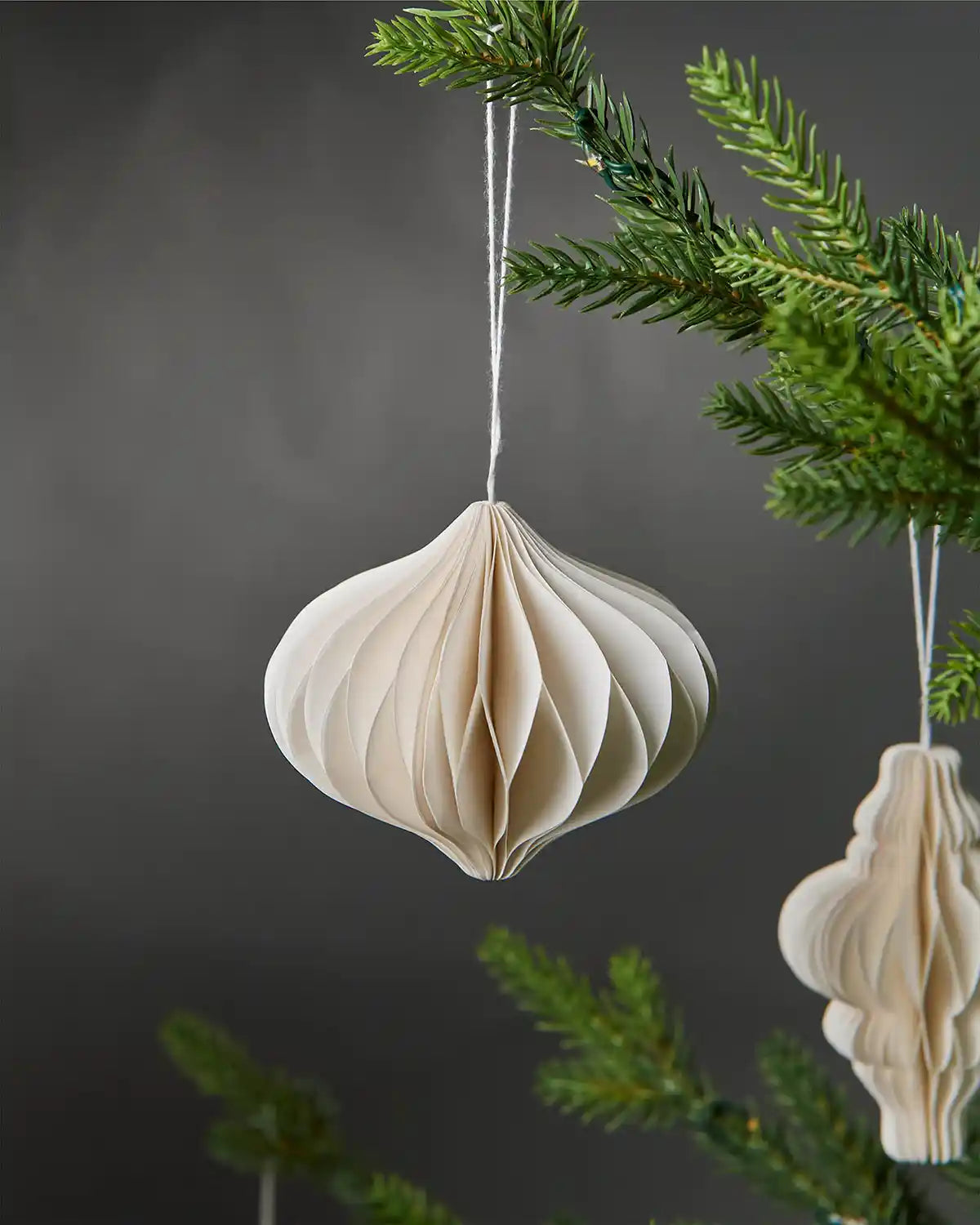 Close-up of an individual 10cm off-white paper decoration hanging on a Christmas tree with another decoration in the background with a grey wall