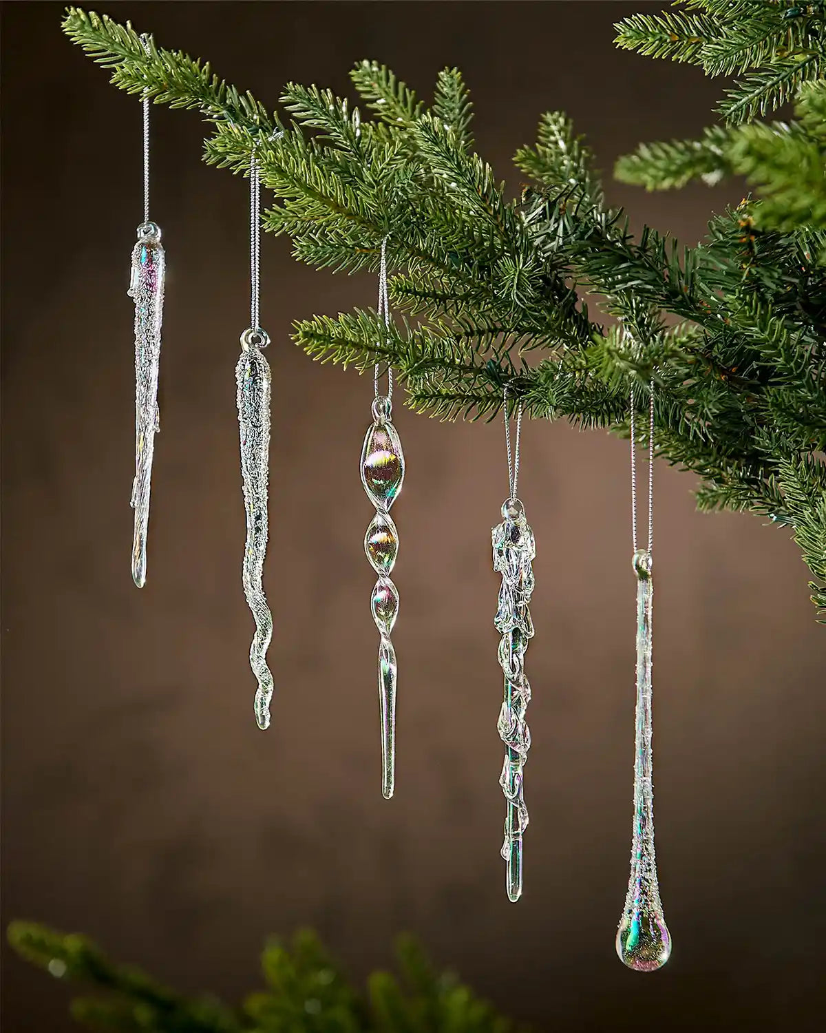 Iridescent icicle ornaments hanging on a Christmas tree branch against a brown background