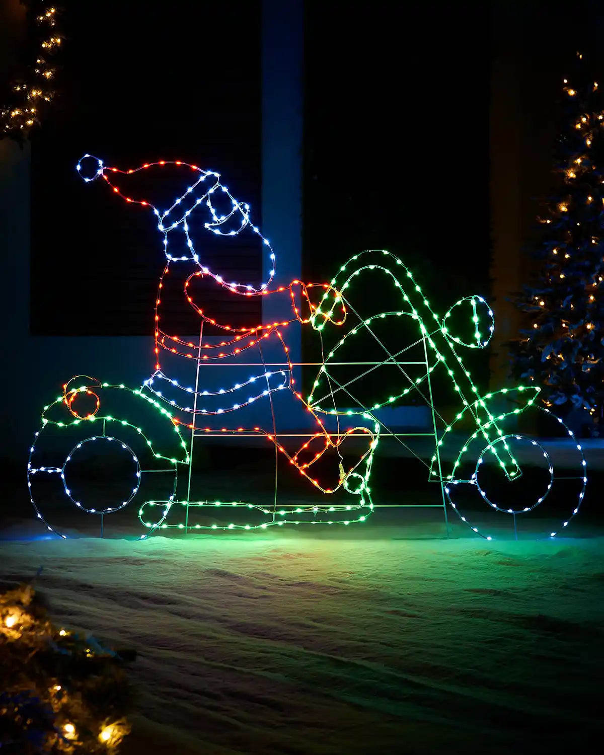 LED Red, green blue and Ice white lights shaped as Santa on a motor bike on a snowy garden floor with a decorated Christmas tree in the background