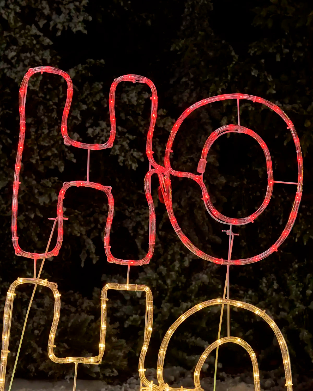 LED lighted wire sign spelling 'HO HO' in red and warm yellow/gold tones, photographed outdoors at night against a dark foliage background.