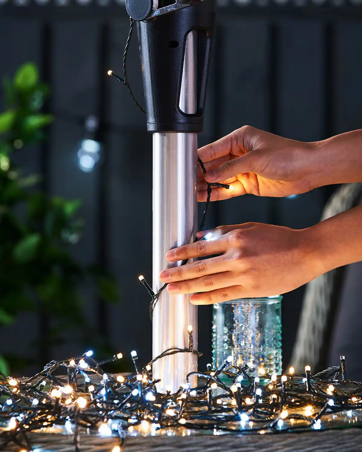 Lady adjusting warm white and ice white string lights on an umbrella pole with a grey fence in background