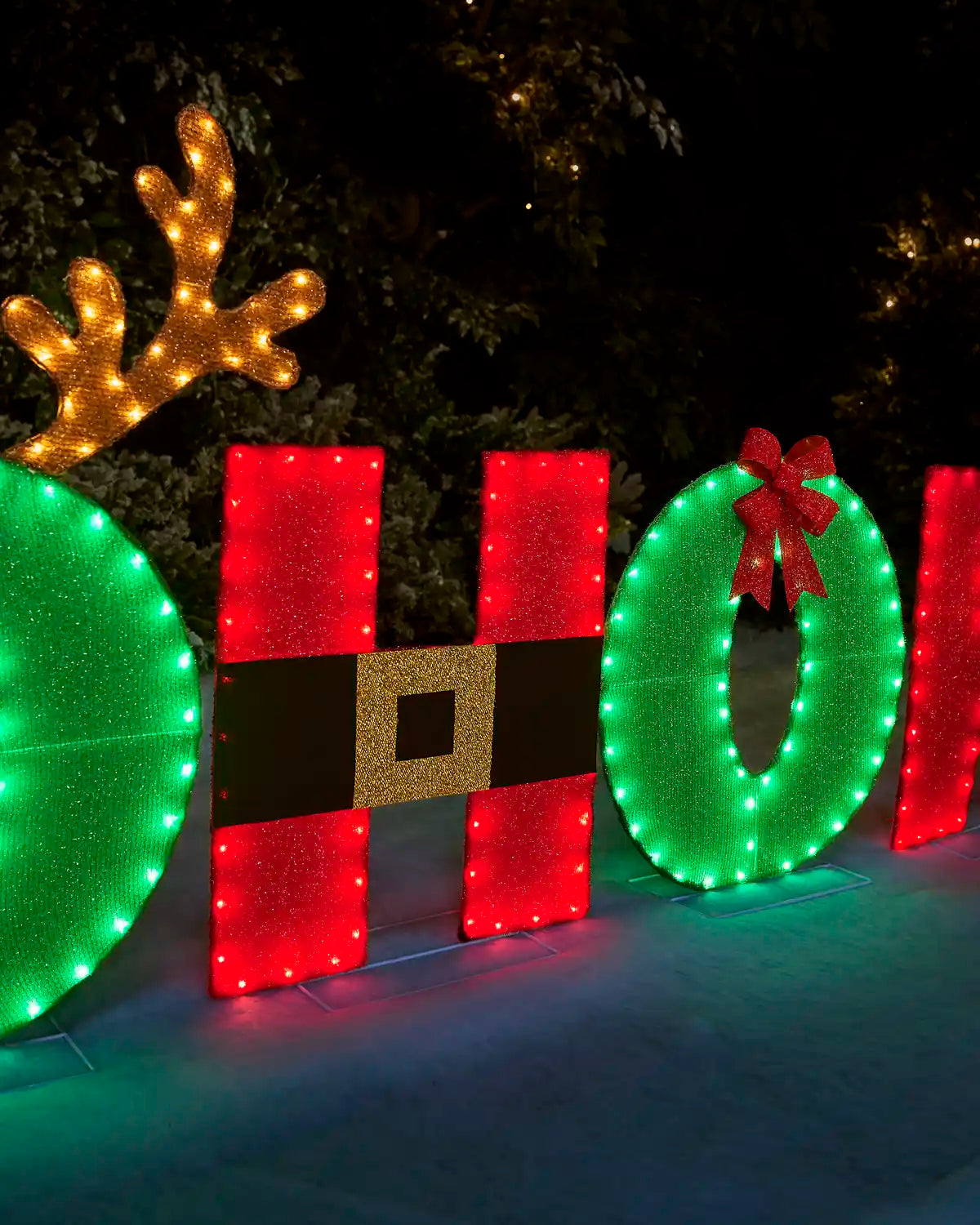 Large holiday LED light decorations spelling out 'HO' with a green wreath, red and black Santa-style belt motif, gold reindeer antlers, and a red bow, photographed at night outdoors.