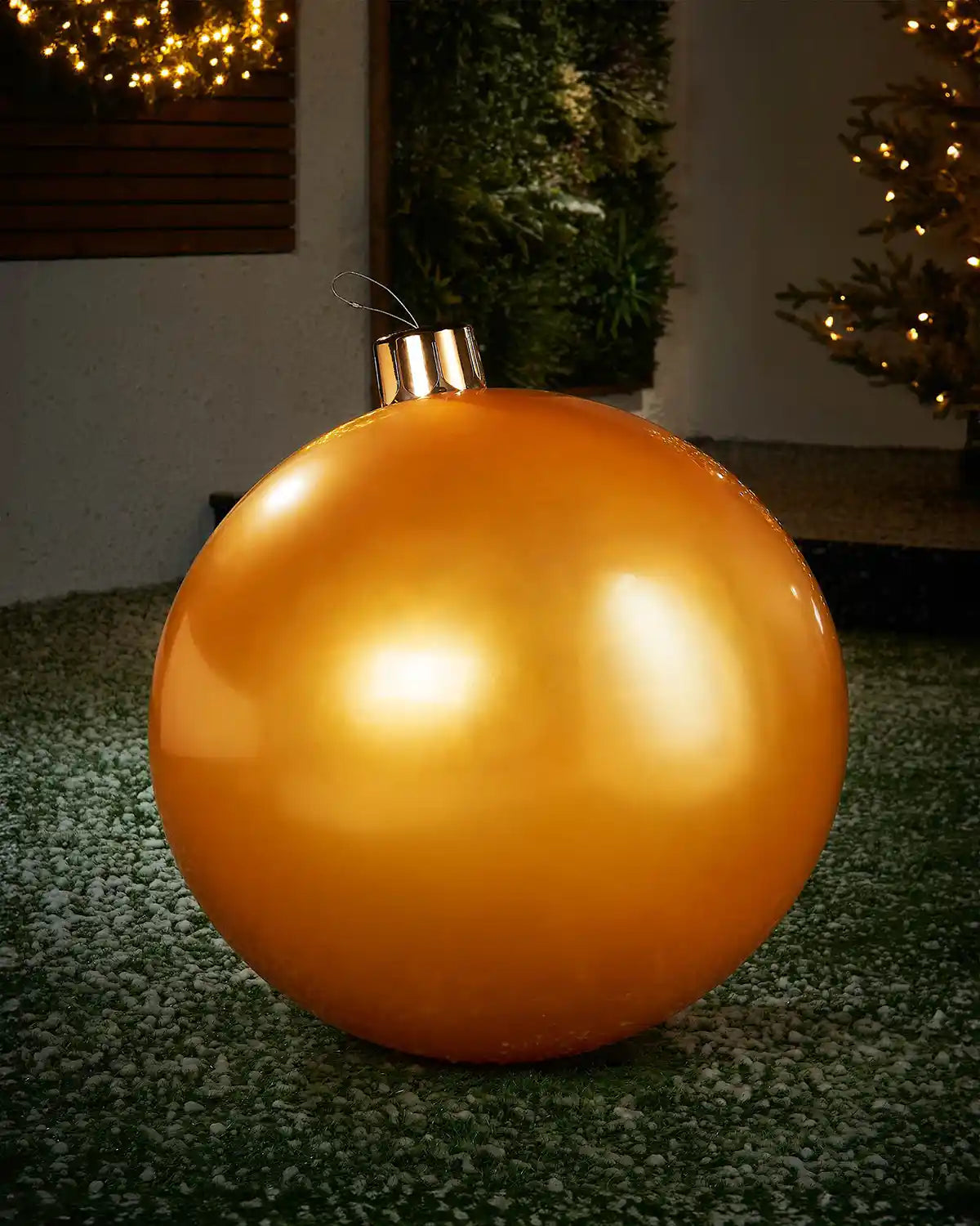 Large sized gold inflatable bauble on a snowy garden floor with a decorated Christmas tree and wreath in background 
