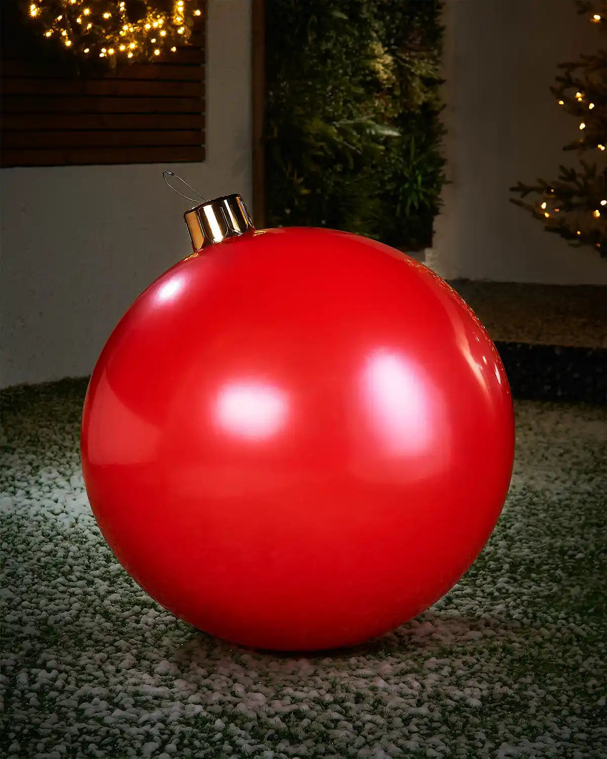 Large sized red inflatable bauble on a snowy garden floor with a decorated Christmas tree and wreath in background 
