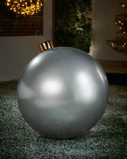 Large sized silver inflatable bauble on a snowy garden floor with a decorated Christmas tree and wreath in background 