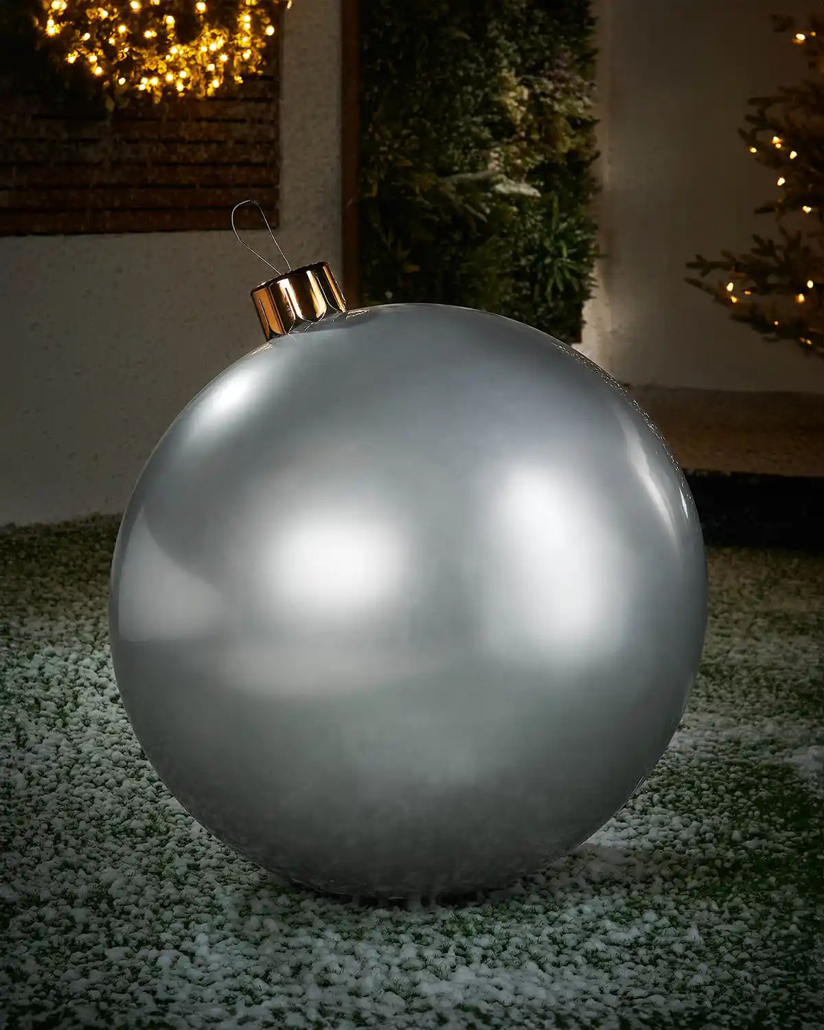 Large sized silver inflatable bauble on a snowy garden floor with a decorated Christmas tree and wreath in background 