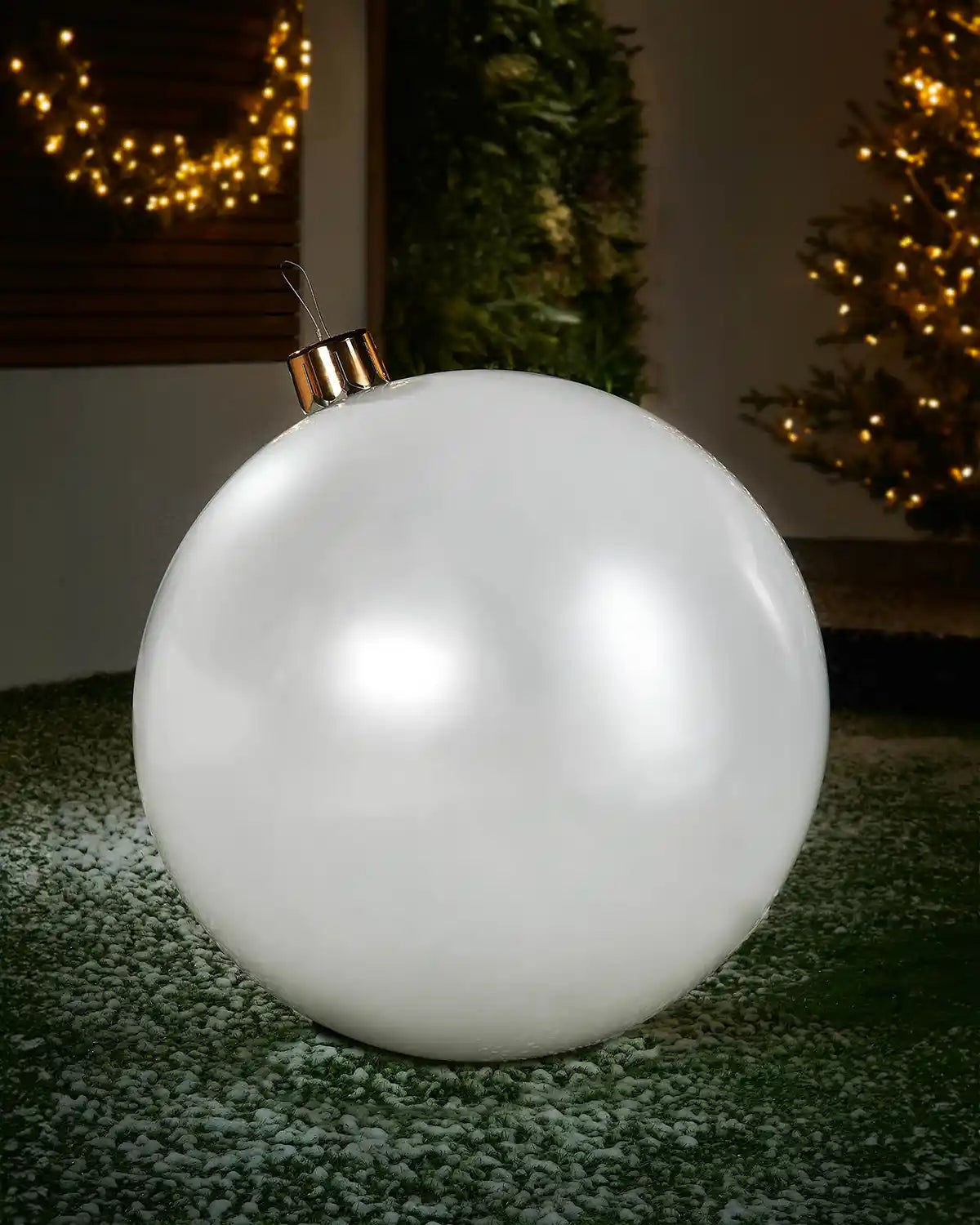 Large sized white inflatable bauble on a snowy garden floor with a decorated Christmas tree and wreath in background 

