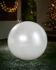 Large sized white inflatable bauble on a snowy garden floor with a decorated Christmas tree and wreath in background 
