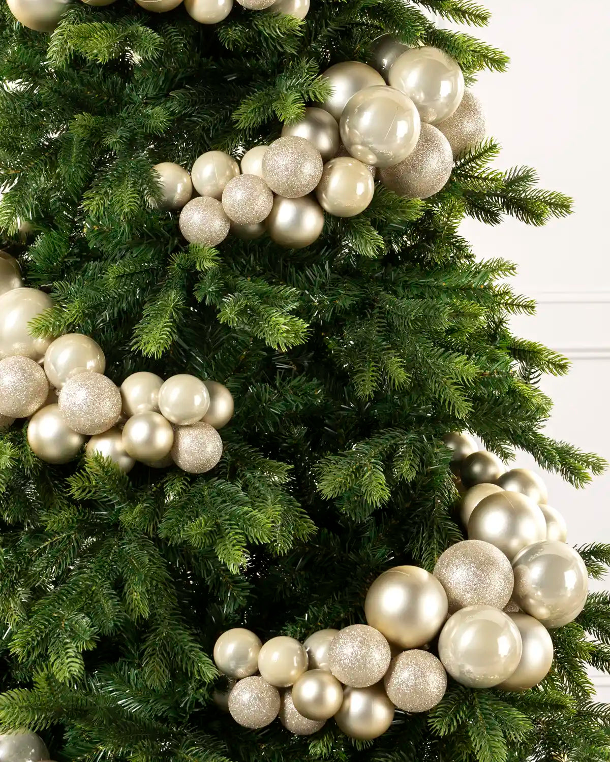 Latte-colored decorative ornaments with both matte and glitter finishes, arranged on a green artificial Christmas tree against a light neutral background.