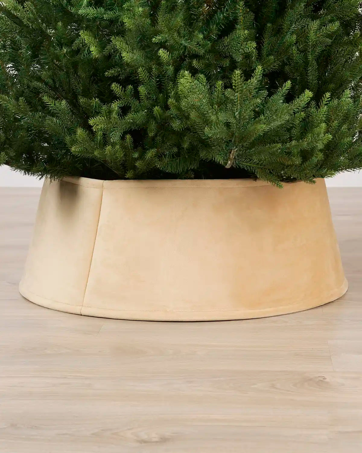 Latte coloured tree skirt holding a green Christmas tree, shown on a light wood floor.