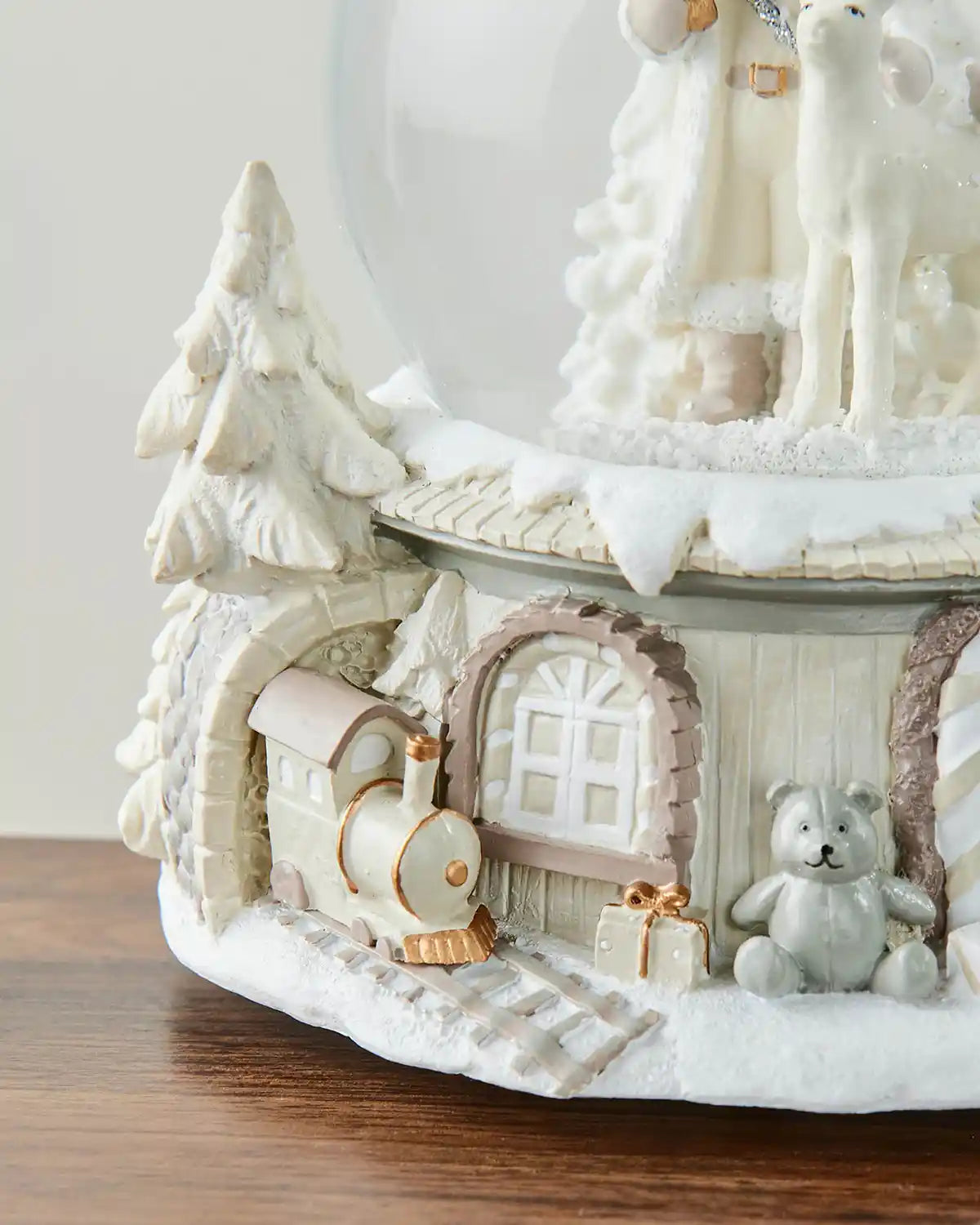Left side close-up of Santa snow globe featuring cream/gold train appearing out of tunnel and silver teddy bear on dark wood surface in front of white wall