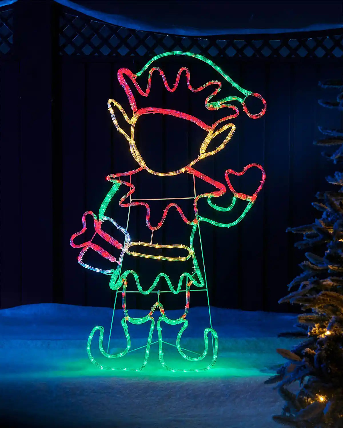 Light up LED elf with red, white, green and warm white colours holding a present on snowy grass with a fence in background against a dark winter sky