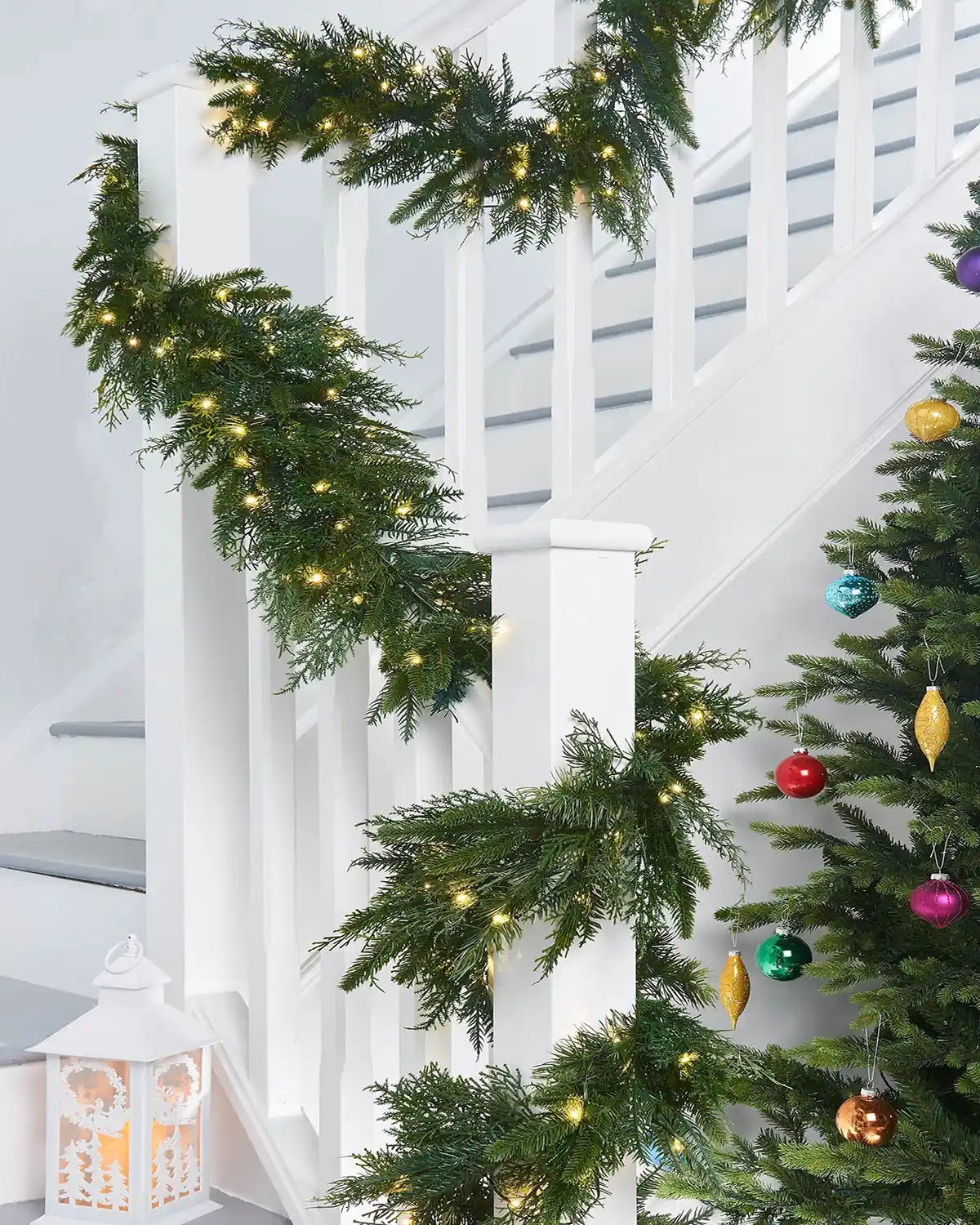 Long view Pre-Lit Natural Fir Garland, 18 ft on a staircase wooden white banister, multi coloured baubles on a Christmas Tree